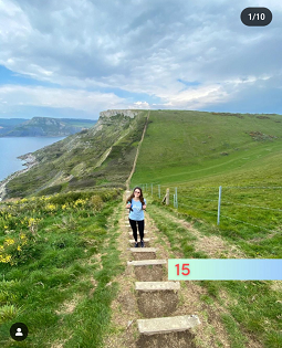 A white female with long brown hair smiles at the camera, standing on wooden steps on a coastal path. 